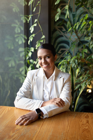 Cheerful pretty young black woman in white suit with smart watch sits at table in cafe with plants...