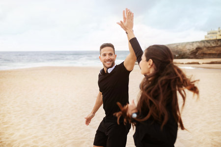Smiling man and woman running in morning on beach near ocean and giving high five, enjoying outdoor workoutの写真素材