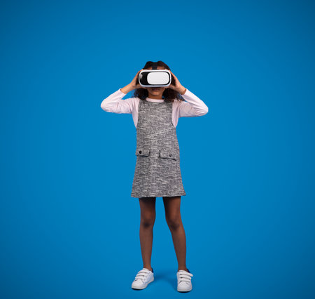 Smiling black little girl in VR glasses plays online game, has fun isolated on blue backgroundの写真素材