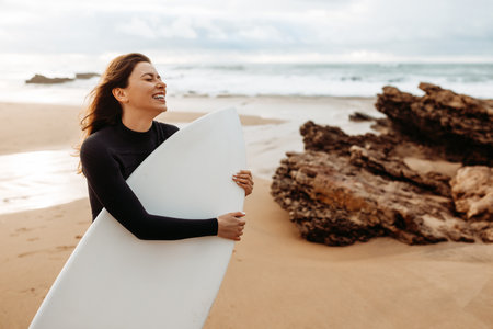 Portrait of excited young surfer woman on the beach holding her surfboard, posing and smiling, enjoying water sportの写真素材