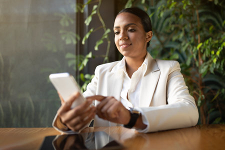 Serious young black lady in white suit chatting on phone, at table with tablet with blank screen in cafeの写真素材