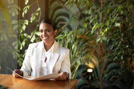 Cheerful young black woman in white suit sitting at table, reading menu in eco cafe with plants interiorの写真素材