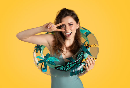Happy woman in swimsuit holding inflatable ring and showing peace sign near eye, posing isolated on yellow backgroundの写真素材