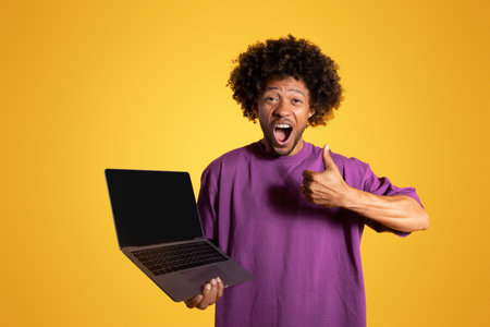 Cheerful excited adult african american curly man show thumb up and laptop with blank screenの写真素材
