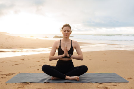 Young calm woman in sportswear practicing yoga, meditating with closed eyes on beach near ocean, copy spaceの写真素材