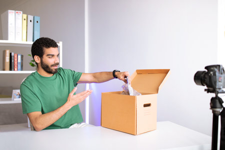 Smiling young middle eastern guy blogger at table unpacks cardboard box making video reviewの写真素材