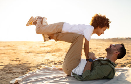 Young father and his son sharing a special moment on the coastline, man lying on plaid and raising the boy on his feetの写真素材