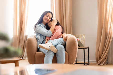 Korean Mother Embracing Baby Daughter Sitting In Armchair At Homeの写真素材