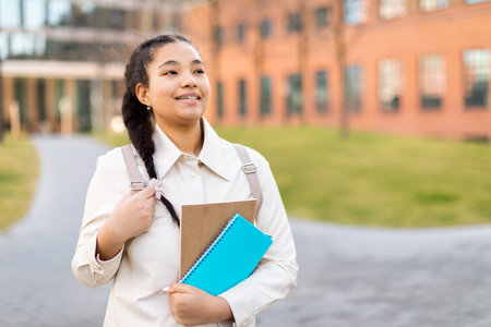 Portrait of positive student lady standing outdoors, teen with workbooks and backpack walking at college campusの写真素材
