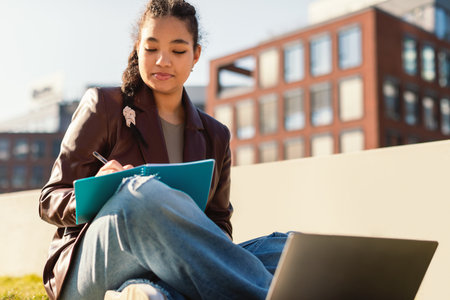 Online education concept. Lady mixed race student studying outdoors with laptop, taking notes during remote lessonの写真素材