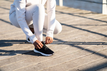 Unrecognizable young black woman in sportswear tying her shoelaces before jogging outdoors