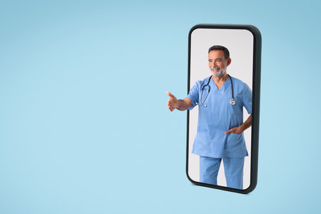 Positive european senior man doctor in uniform greeting empty space with hand on phone screenの写真素材
