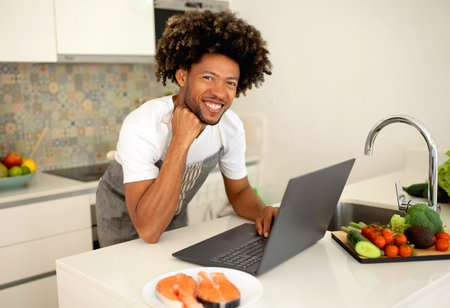 Cheerful Black Man Cooks and Uses Laptop in Modern Kitchenの写真素材