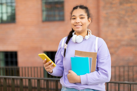 Happy mixed race teen lady with backpack, books and smartphone standing in college campus walking outdoors after classesの写真素材