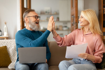 Senior Couple Giving High Five Using Laptop Holding Papers Indoorsの写真素材