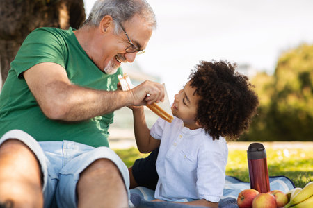 Happy elderly european grandfather and mixed race little boy lie on plaid in park, enjoy free time togetherの写真素材