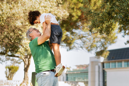 Cheerful elderly european grandfather raises on hands mixed race little boy, have fun in parkの写真素材