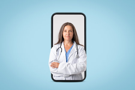 Medical services concept. Old woman doctor in uniform posing with stethoscope and folded arms in giant cellphone screenの写真素材