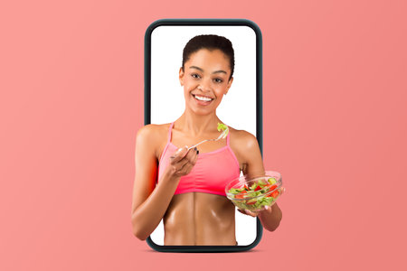 Smiling young african american lady in sports bra eating salad on phone screenの写真素材