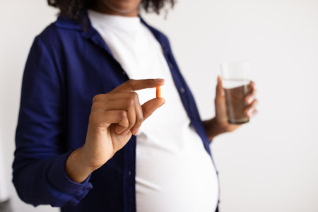Millennial african american pregnant lady with big belly shows capsule pill, glass of waterの写真素材