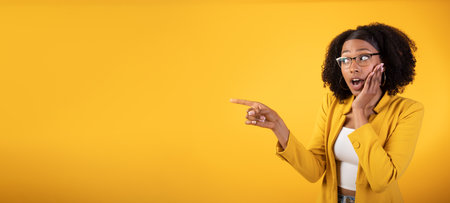 Surprised african american lady pointing aside at blank space, amazed at huge sale over yellow background, panoramaの写真素材