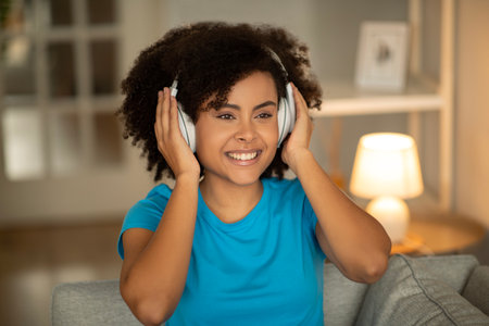 Satisfied happy young black curly woman in wireless headphones enjoys free time alone, listen musicの写真素材