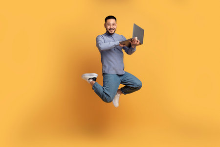 Happy asian man with laptop computer jumping up over yellow backgroundの写真素材