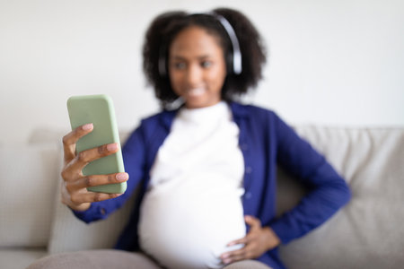 Cheerful millennial black pregnant woman with big belly in headphones looks at phone on sofaの写真素材