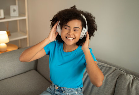 New gadget and audio app. Positive young black curly woman in wireless headphones sits on sofaの写真素材