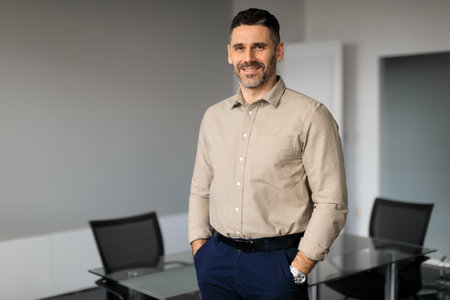 Portrait of confident middle aged businessman posing with hands in pockets in office, looking and...