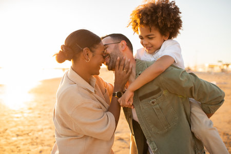 Young happy family spending time outdoors at the beach, spouses father and mother kissing, man piggybacking kid boyの写真素材