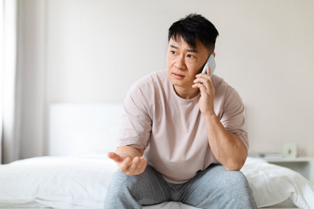 Puzzled chinese man sitting on bed, have phone callの写真素材