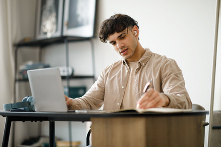 Middle Eastern Guy Using Laptop Websurfing And Taking Notes Indoors