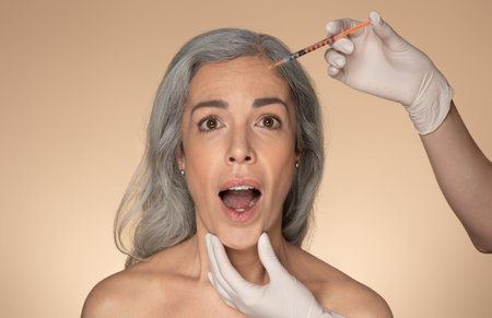 Shocked senior woman receiving beauty shots, using injection medicine to prevent aging, beige studio backgroundの写真素材