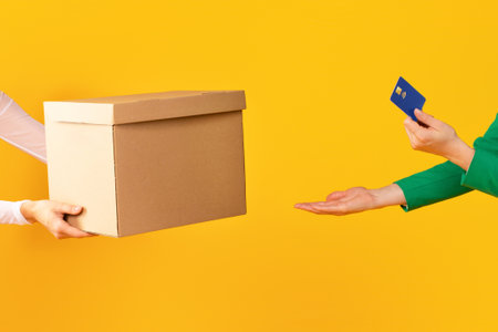 Female hands holding credit card and taking cardboard box from carrier or seller on yellow studio backgroundの写真素材