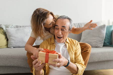 Happy senior woman giving gift box to her husband, congratulating with birthday, sitting on sofa at homeの写真素材