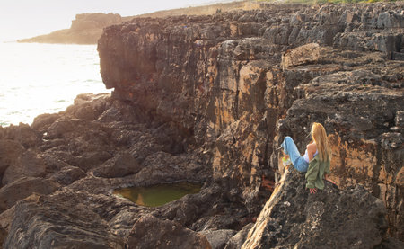 Young woman sitting on rock ledge and looking at ocean bayの写真素材