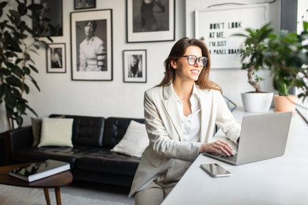 Glad european businesswoman in suit using laptop computer, sitting and looking at window in office interior, free spaceの写真素材