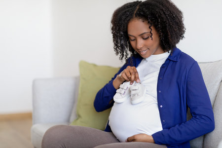Glad curly young black woman in domestic clothes with big belly enjoy pregnancy, hold small shoesの写真素材