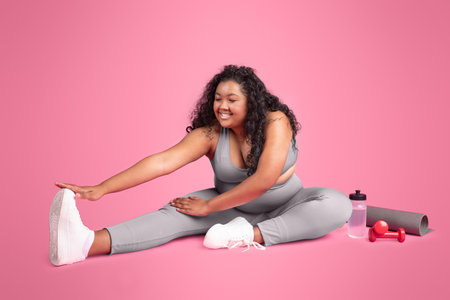 Happy black plus size woman stretching her legs, exercising during workout on pink studio background, free spaceの写真素材