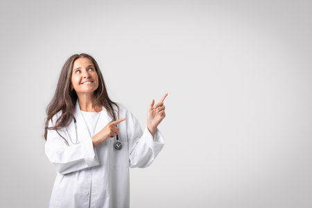 Smiling senior woman doctor in medical workwear pointing at copy space for advertisement, grey background, web-bannerの写真素材