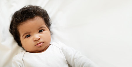 Portrait of cheerful curly sweet black little child boy or girl in clothes lies on white bed in...