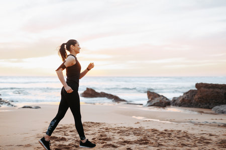 Cheerful slim adult european lady in sportswear race walking, enjoys workout in morning on sea beachの写真素材