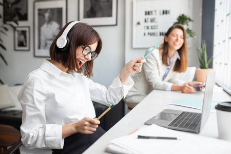 Joyful lady office manager in headphones enjoying music and singing while working in office with colleague on backgroundの写真素材