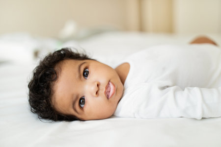 Happy cute sweet black little child lies on white bed in bedroom, kid rest and relax, close up