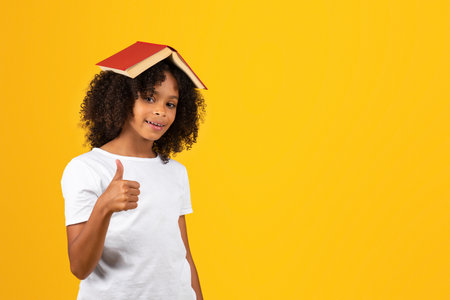 Smiling adolescent curly african american girl in white t-shirt with book on head show thumb upの写真素材