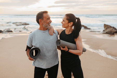 Smiling mature european couple in sportswear with mats enjoy workout together in morning on sea beachの写真素材