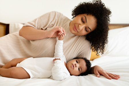 Parenthood and family. Smiling black millennial mother plays with little baby on white bed, enjoys...