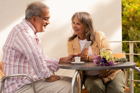 Cheerful senior caucasian lady with bouquet shows phone to husband, drink coffee, sit at table in...