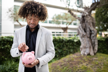 Cheerful black middle aged man putting coin in piggy bank, for dream or new startup, standing outdoors, copy spaceの写真素材
